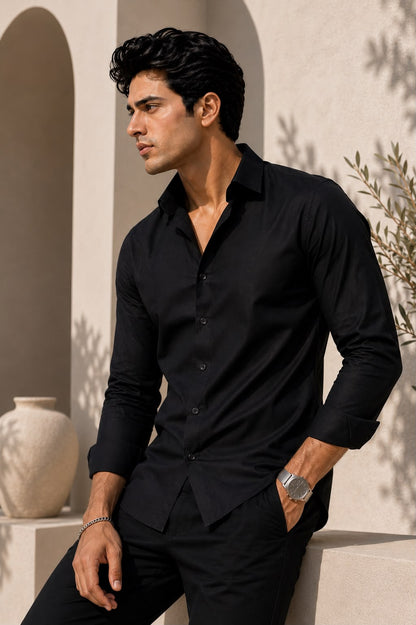 A man wearing a black long-sleeve button-up shirt and black trousers, leaning against a beige stone wall with arches and shadows.