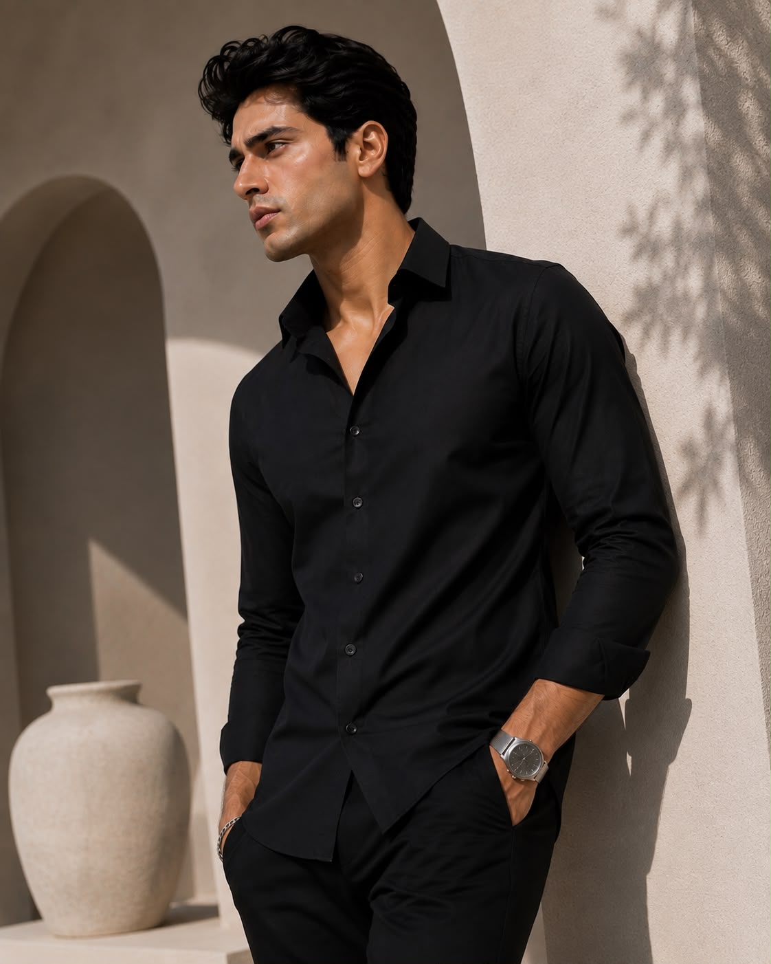 A man wearing a black long-sleeve button-down shirt and matching black trousers, standing in a sunlit architectural setting with beige walls and a large white vase.
