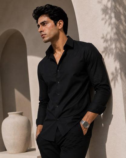 A man wearing a black long-sleeve button-down shirt and matching black trousers, standing in a sunlit architectural setting with beige walls and a large white vase.